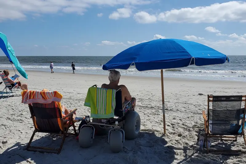A man in a beach wheelchair with wide balloon tires under a blue umbrella, facing the ocean on a Hilton Head beach