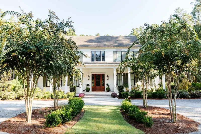 A white house with a columned porch at the end of a walk lined with crape myrtles
