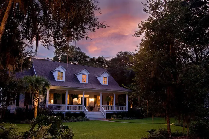 A house with three dormers and a lit front porch under oaks at dusk