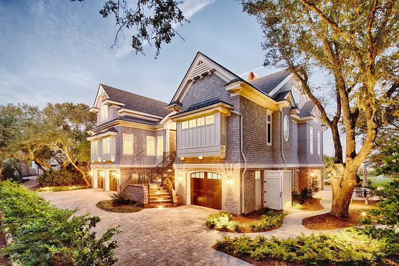 A shingle-sided house on a raised foundation lit up at dusk beside a live oak