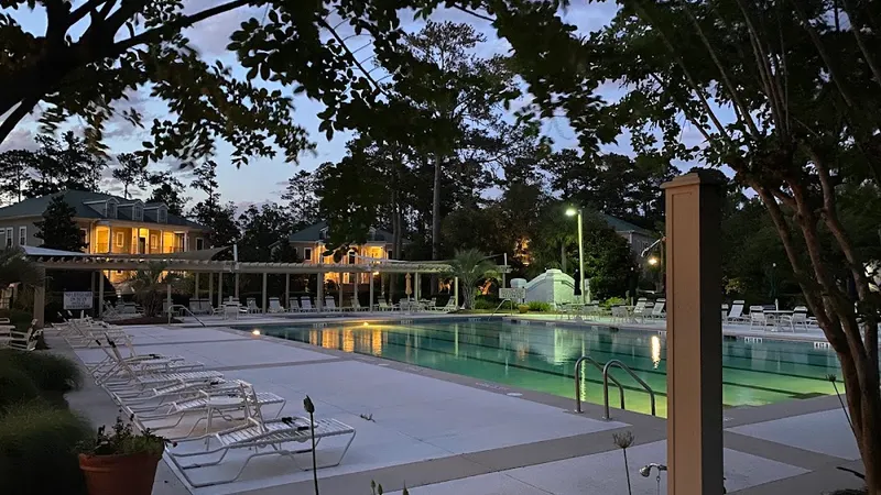 A swimming pool with lounge chairs along its deck under the trees at dusk