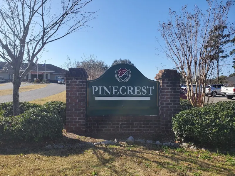A green Pinecrest sign set in a brick wall, with a low building and parked cars behind it