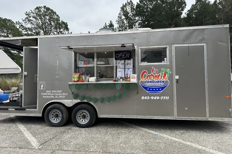 A grey food trailer with its serving window open on a sunny afternoon, parked on gravel under pine trees