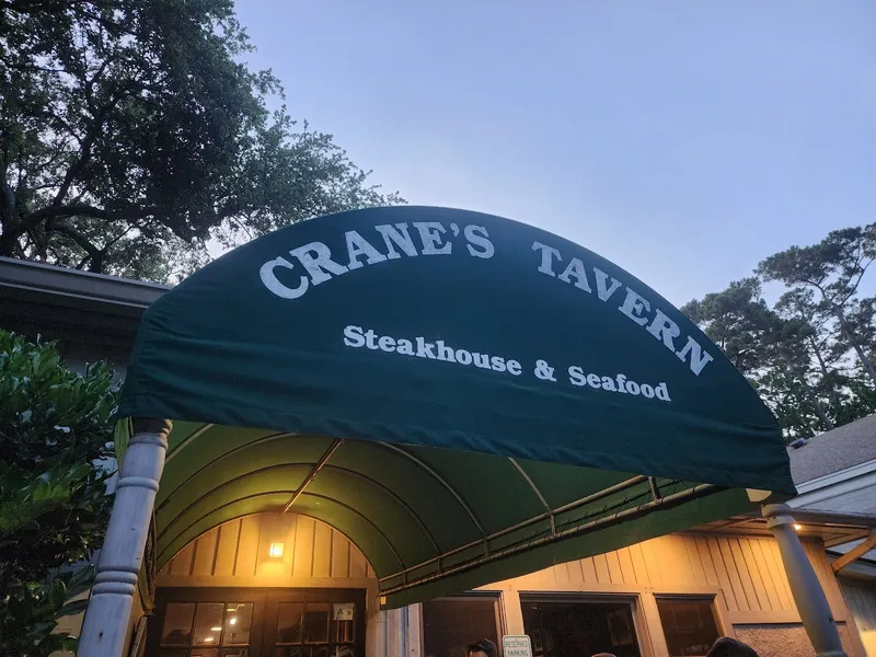 The green awning at the entrance to Crane's Tavern, reading Steakhouse & Seafood, under live oaks at dusk