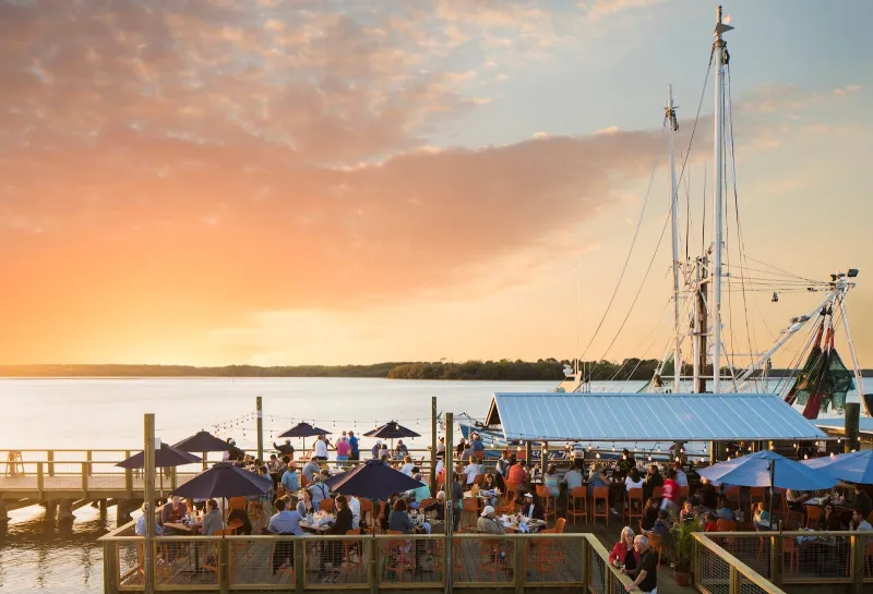 Diners on the covered deck at Hudson’s Seafood House on the Docks at sunset, with a shrimp boat’s masts behind the dock