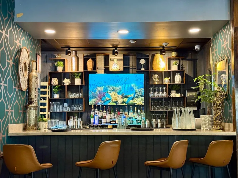 The bar at Masala Mantra Indian Bistro with teal patterned walls, leather stools and a lit shelf of bottles