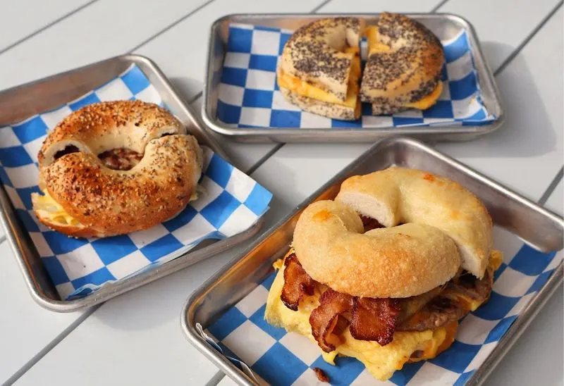 Three bagel breakfast sandwiches in checkered paper trays at Nosh NY Bagels, one with bacon, egg and sausage on a cheese bagel