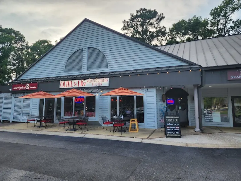 The gray shingled storefront of Olive & Fig Mediterranean Kitchen at Moss Creek Village, with orange umbrellas over outdoor tables