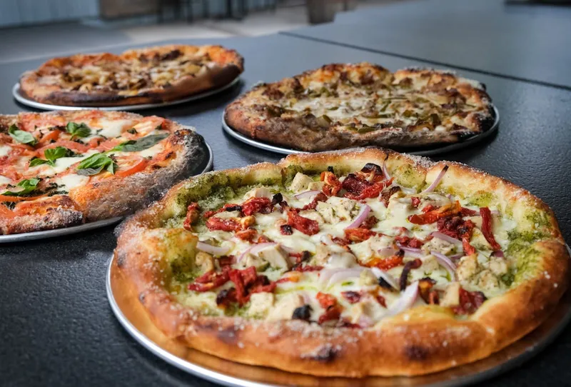 Several thin, charred-crust pizzas on pans, including a pesto pizza with sun-dried tomatoes and a margherita