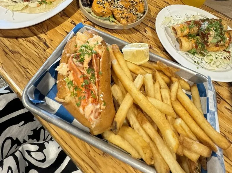 A Slapfish lobster roll and fries in a checkered tray, with tacos and shrimp on plates behind it