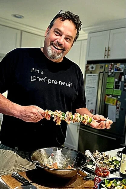 Chef Chris Litts of The Curated Chefs holding a grilled skewer over a wok in a home kitchen