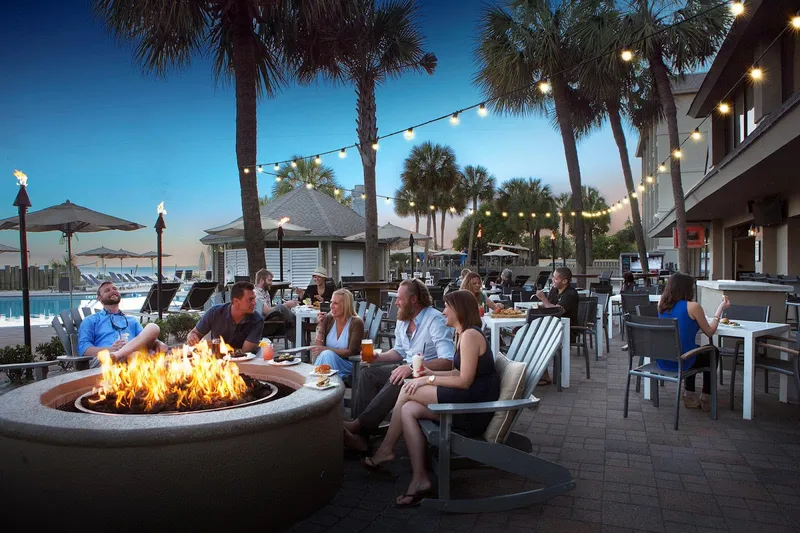 Guests around a fire pit on the string-lit patio at The Porch, with palms and the Beach House pool behind