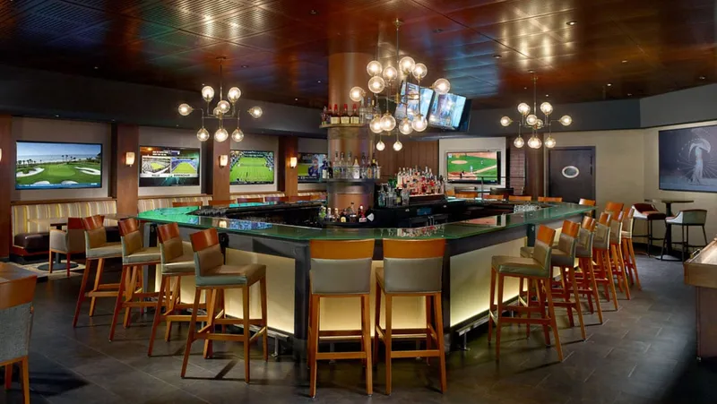 The bar at XO Sports & Spirits, with barstools around a lit island bar and TVs showing golf and other sports