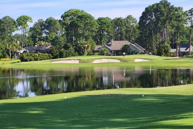 A green guarded by bunkers across a lagoon at Oyster Reef Golf Club, with homes among the pines behind
