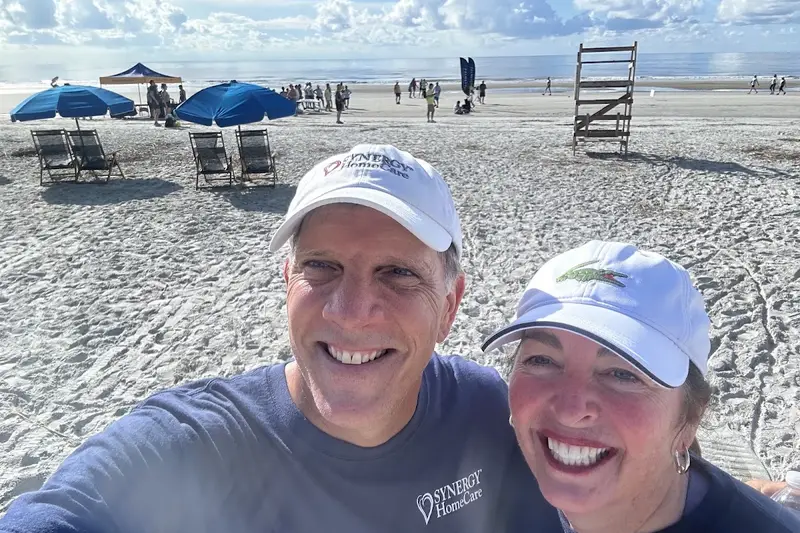 Two SYNERGY HomeCare team members in company caps, smiling on the beach