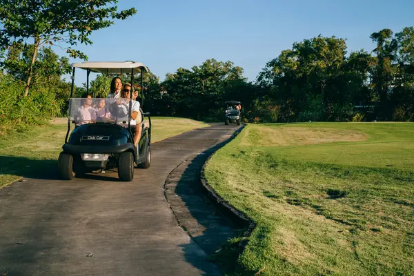 A white four-seat golf cart with passengers on a cart path beside a fairway