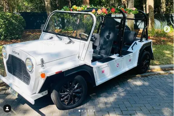 A white Moke from Slow Rides decorated with a garland of flowers, parked under the trees