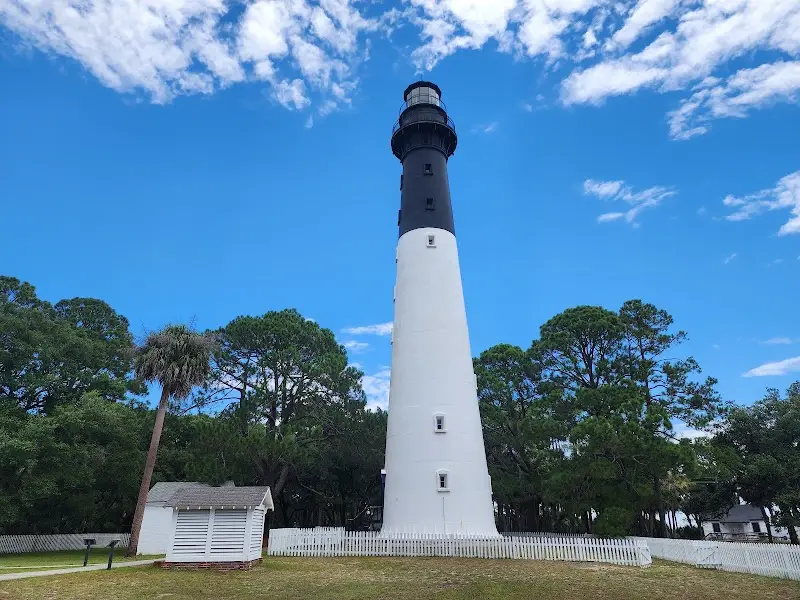 The Hunting Island Lighthouse, white below and black at the top, with a small white outbuilding and a picket fence