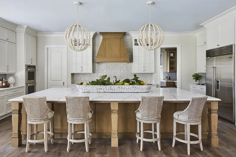 A white kitchen with a wood range hood, a long island and woven bar stools
