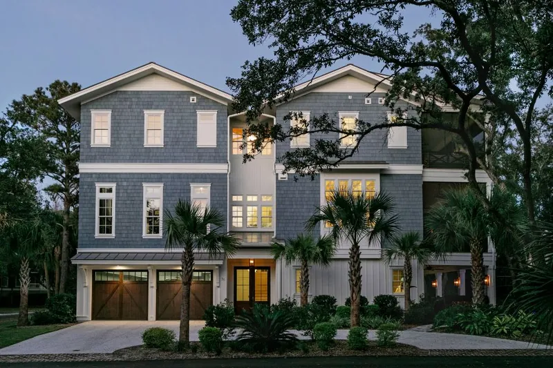 A three-story shingle-sided house with lit windows and palms out front at dusk