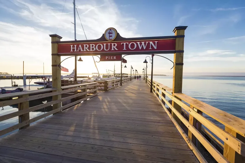 The Harbour Town Pier sign over the wooden walkway of the covered pier at sunrise, with the marina on one side and Calibogue Sound on the other