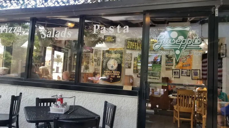 The front windows of Giuseppi’s Pizza in Sea Pines, lettered Pizza, Salads, Pasta, with patio tables outside