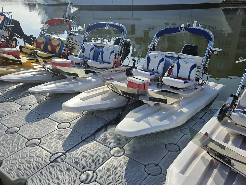 A row of small two-seat catamarans with blue canopies tied up on a floating dock, a white tour boat behind them