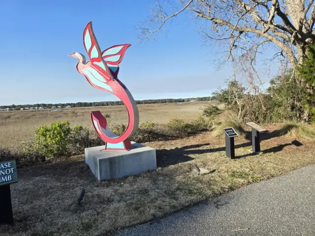 A red and blue bird sculpture on a plinth beside the path, with the Broad Creek marsh behind it