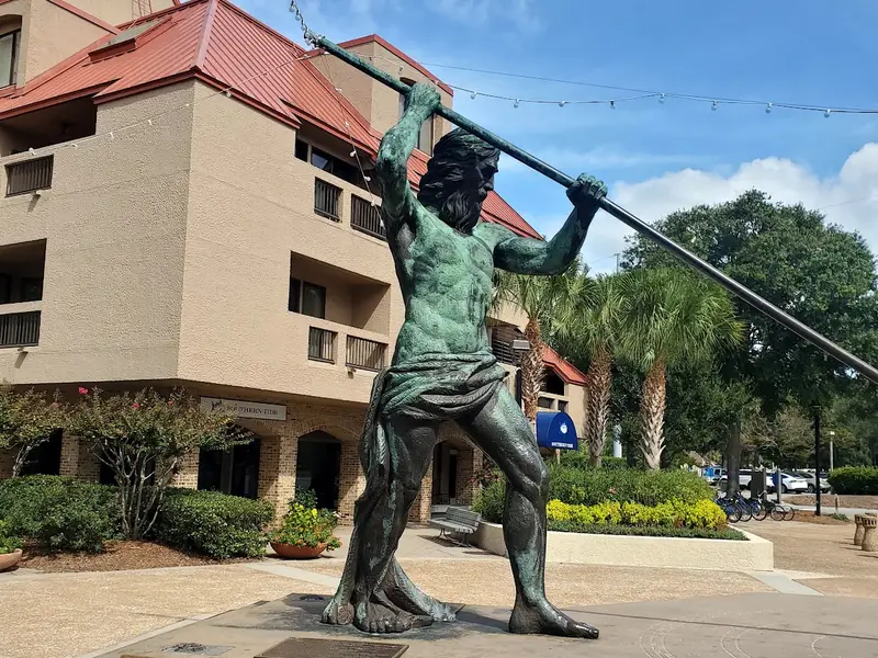 The bronze King Neptune statue on its stepped plaza at Shelter Cove Harbour, trident raised overhead against a blue sky