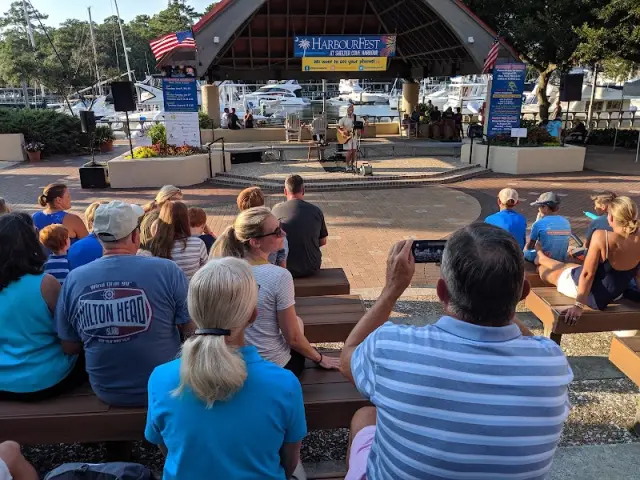 An evening audience on benches in front of the HarbourFest stage at Shelter Cove Harbour, with boats behind it