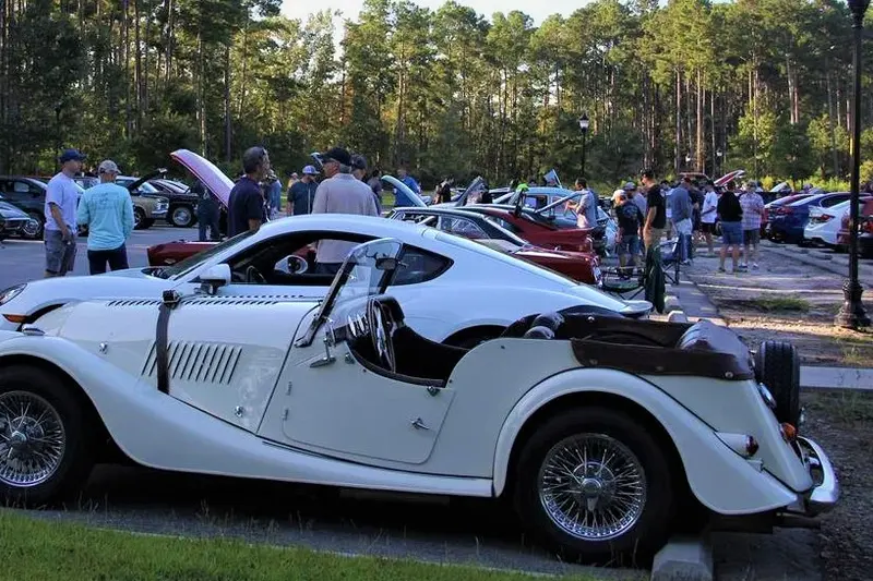 A white neoclassic roadster and a crowd walking a lot of parked cars at the first-Saturday morning car meet