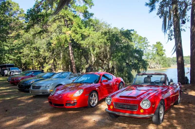 Red sports cars and other club cars parked under live oaks beside the water on a Lowcountry Oyster & Motorcar Driving Society outing