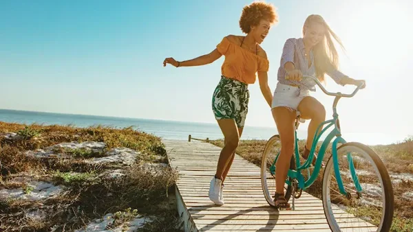 Two women on a beach boardwalk, one riding a teal cruiser bike