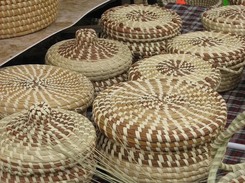 Stacks of coiled sweetgrass baskets with lids on a checked tablecloth, with loose sweetgrass in front