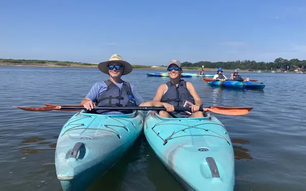 Two smiling paddlers in turquoise kayaks on Broad Creek, with more of the group behind them