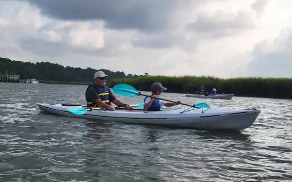 An adult and a child paddling a white tandem kayak past the marsh grass on Broad Creek